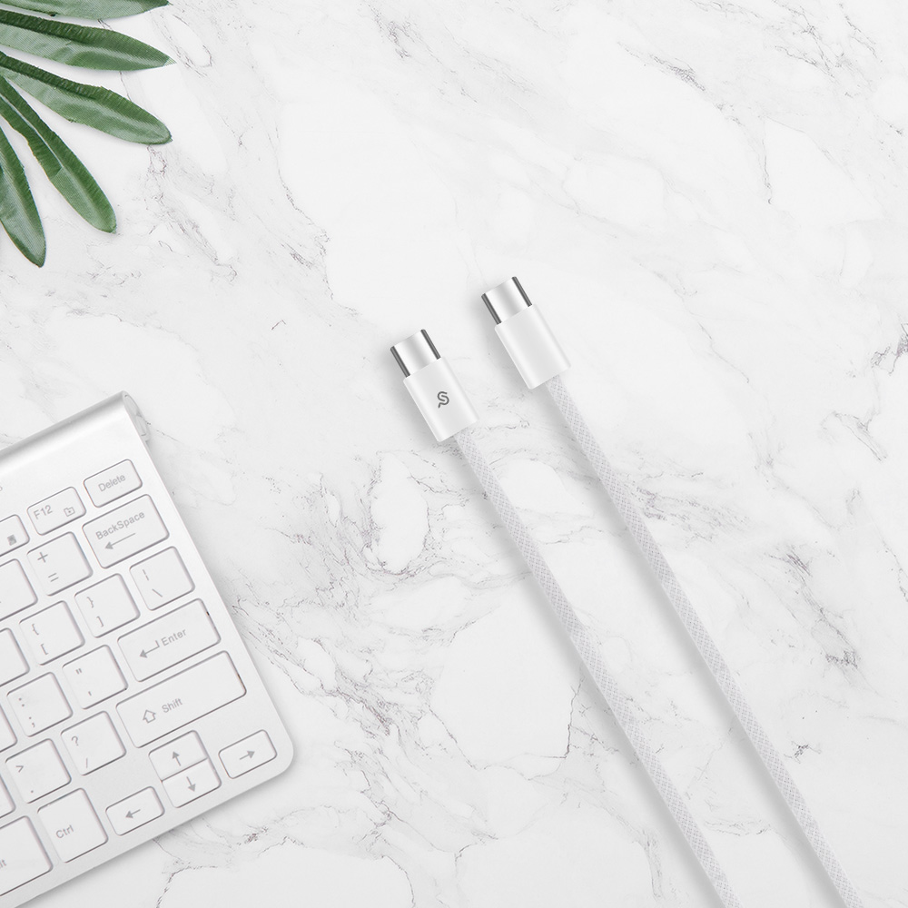 White braided USB-C to USB-C cable laid on a white marble surface, alongside a white keyboard and green plant leaves