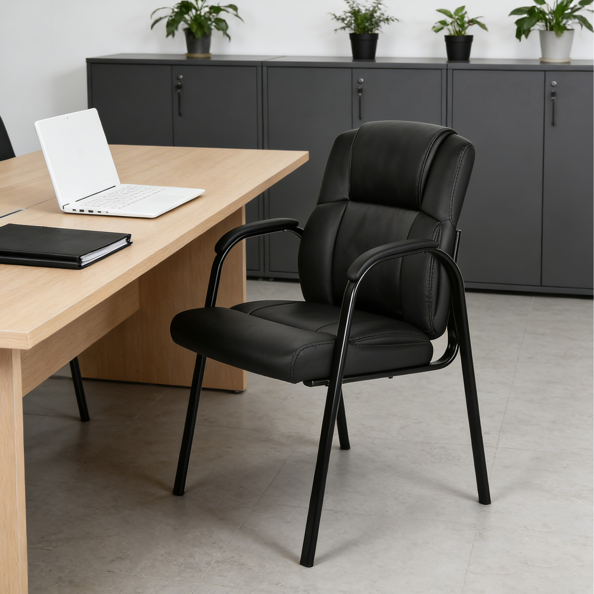 Black PU leather high back guest chair positioned beside a light wood office desk and in front of the grey storage cabinets